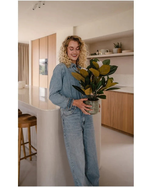 Una mujer con cabello rubio y rizado, vestida con ropa vaquera, sonríe en una cocina moderna con la planta de magnolia artificial en una maceta de 46 cm en sus manos, rodeada de armarios y estantes de madera: una opción ideal para cualquiera que busque plantas de bajo mantenimiento para realzar su elegante interior.
