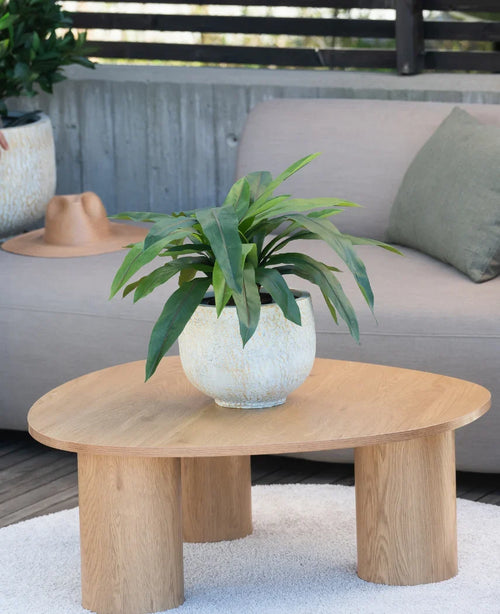 Una planta artificial Bird's Deluxe de 60 cm se encuentra sobre una mesa de centro redonda de madera frente a un sofá beige, decorada con un cojín verde y un sombrero marrón que cuelga sobre el reposabrazos.