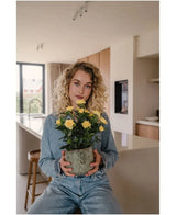 Una mujer con cabello rubio rizado, vestida con ropa vaquera, sentada en una cocina moderna con la Planta Artificial Mini Rose UV 35 cm amarilla en sus manos y mira directamente a la cámara.