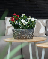 Una planta artificial de jardín rosa de 35 cm color crema UV se encuentra sobre una mesa redonda de madera frente a un sofá beige con cojines estampados.