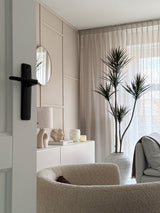 Un salón moderno y minimalista con un sillón con textura beige, una planta artificial Dracaena Fragnans de 180 cm, un aparador blanco con decoración, un espejo redondo y cortinas transparentes que filtran la suave luz del día a través de la ventana.