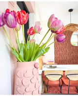 Un jarrón con motivos de corazones rosas sostiene una flor artificial de tulipán francés de 60 cm de color violeta sobre una mesa de comedor con sillas con respaldo tejido. Al fondo, un espejo redondo y una pared con motivos marrones contribuyen a crear un interior acogedor y moderno.