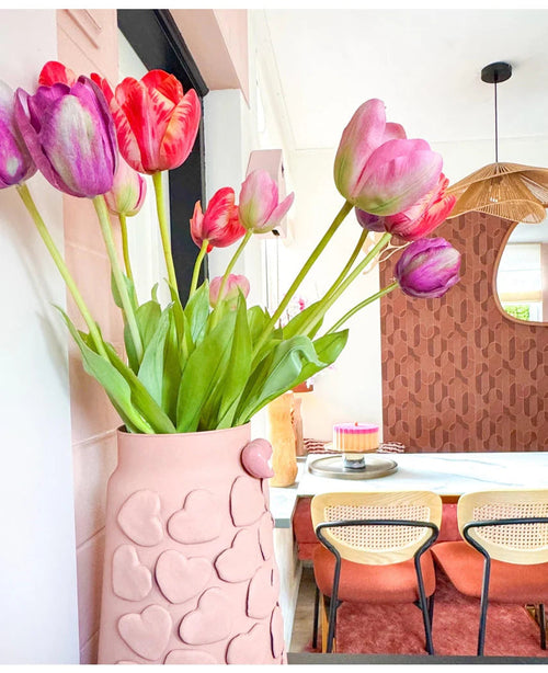Un jarrón con motivos de corazones rosas sostiene una flor artificial de tulipán francés de 60 cm de color violeta sobre una mesa de comedor con sillas con respaldo tejido. Al fondo, un espejo redondo y una pared con motivos marrones contribuyen a crear un interior acogedor y moderno.