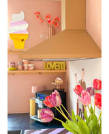 Una cocina rosa pastel con una flor artificial de tulipán francés de 60 cm de color violeta en primer plano, un plato LOVETTI amarillo en el estante, tazas rosas, un arte mural de un cono de helado y un mural minimalista de dos personas en la pared.