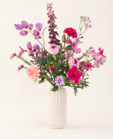 Un jarrón con textura blanca contiene el ramo de flores artificiales de seda Purple Dawn en tonos rosa, morado y lavanda con follaje verde, sobre un fondo claro y neutro.
