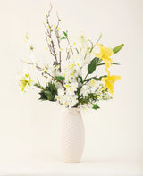 Un jarrón con estructura blanca muestra la elegancia del ramo de flores artificiales de seda Sunkissed Ivory, con lirios amarillos, flores blancas y hojas verdes sobre un fondo liso y claro.