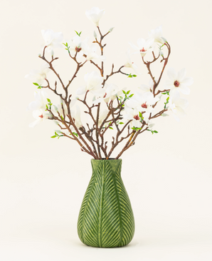 Un jarrón verde muestra el ramo de flores artificiales de seda Magnolia blanco, con varias ramas de magnolia blanca con pequeñas hojas verdes, sobre un fondo liso y claro.