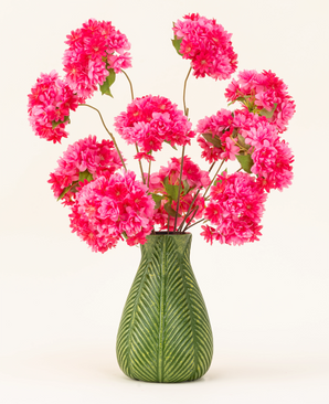 Un jarrón verde con motivo de hojas muestra el ramo de flores artificiales de seda Cherry Blossom Pink (flores de cerezo de seda esponjosas y de color rosa brillante con hojas verdes) sobre un fondo liso y claro.