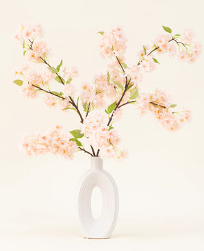Un jarrón de cerámica blanca con una amplia abertura ovalada muestra varios ramo de flores artificiales de seda de hojas verdes, ramas de cerezo en flor de color rosa claro sobre un fondo claro simple.