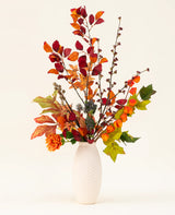 Un jarrón con textura blanca exhibe el ramo de flores artificiales de seda con dijes de castaño, que presenta hojas de otoño, flores de color naranja y follaje verde: un elegante arreglo artificial sobre un fondo liso y claro.