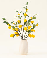 Un jarrón con textura blanca muestra el ramo de flores artificiales de seda Lemons, con hojas verdes y limones amarillos vibrantes sobre un fondo claro.