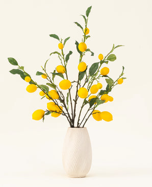 Un jarrón con textura blanca muestra el ramo de flores artificiales de seda Lemons, con hojas verdes y limones amarillos vibrantes sobre un fondo claro.