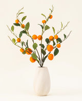 El ramo de flores artificiales de seda mandarina tiene un jarrón con textura blanca y ramas de hojas verdes y mandarinas artificiales de color naranja vibrante, que añaden un toque animado sobre un fondo claro y liso.