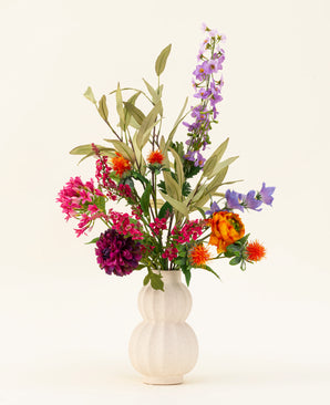 Un jarrón blanco texturizado exhibe el colorido ramo de flores artificiales de seda Svenski, que presenta vibrantes flores artificiales de color púrpura, naranja, rosa y azul con hojas verdes que iluminan cualquier espacio contra un fondo claro simple.