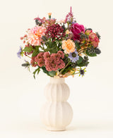 En un jarrón de cerámica blanco y curvo se encuentra el ramo de flores artificiales de seda Pretty Pastel, una colorida mezcla de flores sostenibles como rosas rosadas, dalias, flores violetas y naranjas y vegetación, bellamente dispuestas sobre un fondo claro.