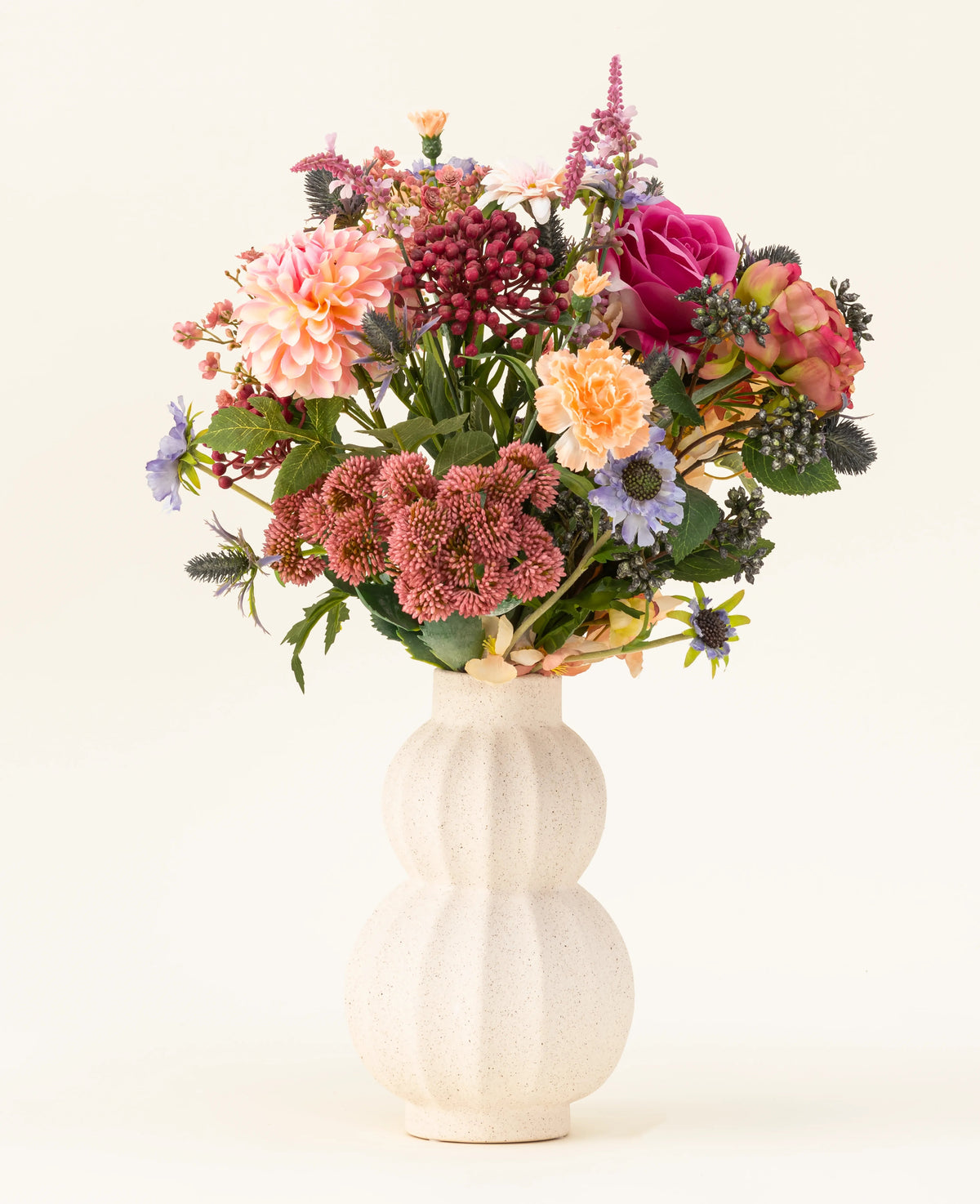 En un jarrón de cerámica blanco y curvo se encuentra el ramo de flores artificiales de seda Pretty Pastel, una colorida mezcla de flores sostenibles como rosas rosadas, dalias, flores violetas y naranjas y vegetación, bellamente dispuestas sobre un fondo claro.
