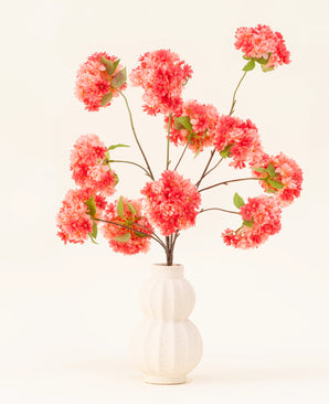 El ramo de flores artificiales de seda Coral en flor de cerezo presenta vibrantes flores de cerezo artificiales de color rosa y coral con hojas verdes, elegantemente dispuestas en un jarrón esculpido en blanco sobre un fondo claro.