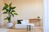 Un salón minimalista con la planta Calocasia artificial de 210 cm, un sofá blanco texturizado con cojines verdes y blancos, dos mesitas auxiliares, una alfombra beige y una mesa consola de madera contra una pared beige claro.