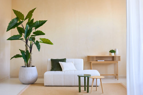 Un salón minimalista con la planta Calocasia artificial de 210 cm, un sofá blanco texturizado con cojines verdes y blancos, dos mesitas auxiliares, una alfombra beige y una mesa consola de madera contra una pared beige claro.