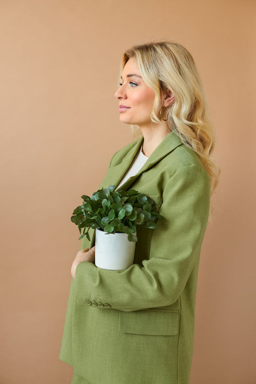 Una mujer de pelo largo y rubio, vestida con una chaqueta verde claro, sostiene contra su pecho la "Planta suculenta artificial de 22 cm en maceta decorativa" y está de lado, frente a un fondo beige liso.