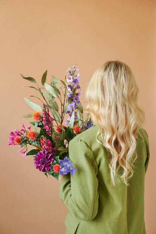 Desde atrás se ve a una persona con cabello rubio largo y ondulado y una chaqueta verde, sosteniendo el colorido ramo de flores artificiales de seda Svenski sobre un fondo beige liso.