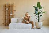 Una mujer vestida de marrón está sentada en un sofá blanco y sonríe con una taza en la mano. Junto a ella se encuentra la planta artificial Strelitzia de 180 cm real touch, que aporta una exuberante vegetación a un salón moderno, teñido de beige y con un mobiliario minimalista.