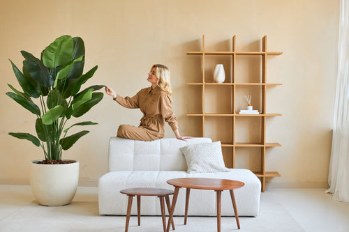 Una mujer con un mono marrón sentada en el respaldo de un sofá blanco y toca la hoja de una planta Strelitzia Artificial Real Touch Deluxe de 180 cm en un salón minimalista con mesas y estanterías de madera.