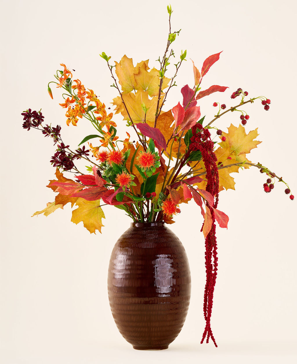 En un jarrón marrón se encuentra el ramo de flores artificiales de seda 'Rustic Glow', con hojas amarillas, naranjas y rojas, frutos rojos, flores naranjas y amarantos rojos largos y colgantes sobre un fondo neutro.