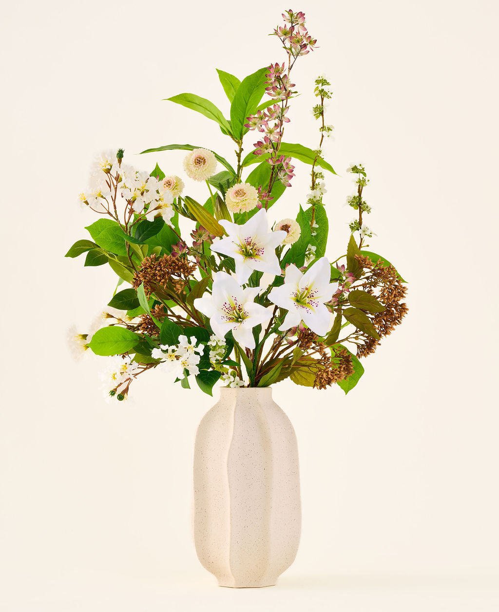 Ramo de flores artificiales de seda Timeless Bloom, compuesto por un ramo de varias azucenas blancas, pequeñas flores blancas y vegetación en un jarrón con estructura de color beige.