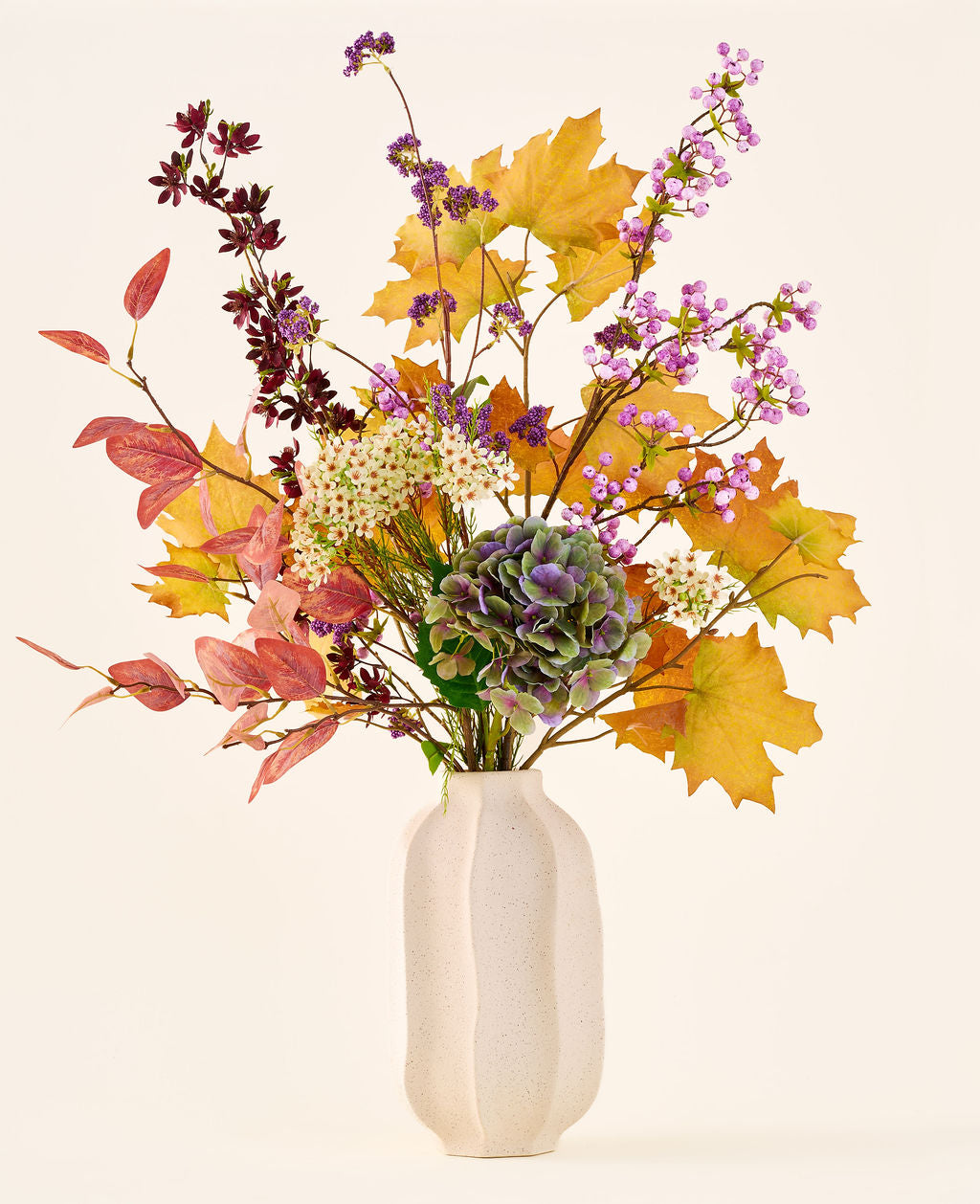 El ramo de flores artificiales de seda 'Wild Amethyst', con hojas otoñales, flores violetas y blancas y hortensias verdes, se presenta en un jarrón de cerámica blanca sobre un fondo liso y claro.