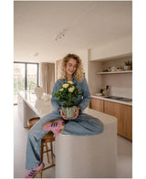 Una mujer con cabello rubio y rizado, vestida con jeans y calcetines a rayas, está sentada sonriendo en la encimera de la cocina mientras sostiene la planta Artificial Gardenia UV de 39 cm, una planta en maceta con flores amarillas. Al fondo se puede ver una cocina moderna con armarios y estanterías de madera.