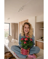 Una mujer sonriente con cabello rubio rizado está sentada con las piernas cruzadas sobre una encimera moderna y luminosa, sosteniendo la planta fucsia Artificial Gardenia UV de 39 cm, una planta en maceta rosa con flores rojas vibrantes. Lleva jeans, una camisa azul y zapatos rosas.