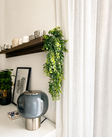Un salón moderno con una lámpara de cristal gris, cuadros enmarcados, una maceta y un estante de madera con tazas y la planta colgante artificial 'Pannenkoek' (61 cm), cuyos zarcillos cuelgan junto a cortinas blancas.