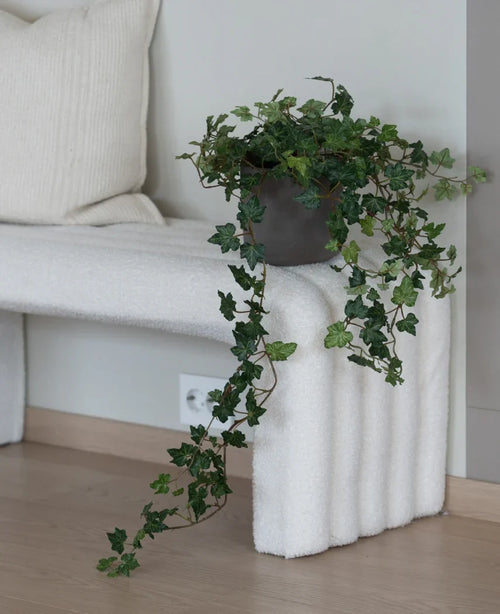 La hiedra artificial Deluxe Green de 90 cm, con sus exuberantes zarcillos colgantes, se encuentra sobre un banco estructurado blanco junto a un cojín de color crema en una habitación minimalista con suelos de madera clara.