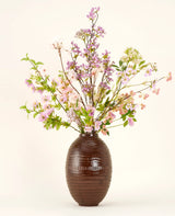 Un jarrón de cerámica marrón muestra el ramo de flores artificiales Silk Lila, un alto y elegante ramo de seda con flores rosas, violetas y lilas con hojas verdes y finas ramas sobre un fondo liso y claro.