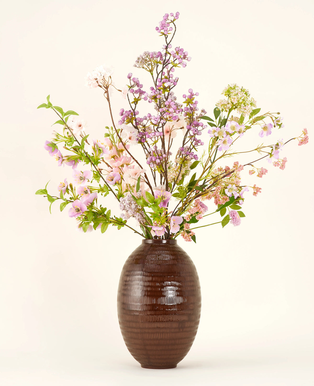 Un jarrón de cerámica marrón muestra el ramo de flores artificiales Silk Lila, un alto y elegante ramo de seda con flores rosas, violetas y lilas con hojas verdes y finas ramas sobre un fondo liso y claro.