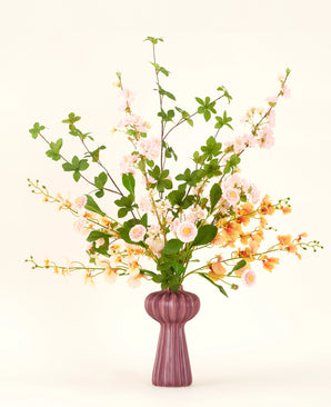 Un ramo de flores artificiales de seda Blossom Harmony está dispuesto en un jarrón malva con flores rosas y duraznos y hojas verdes, sobre un fondo claro simple para una exhibición elegante.