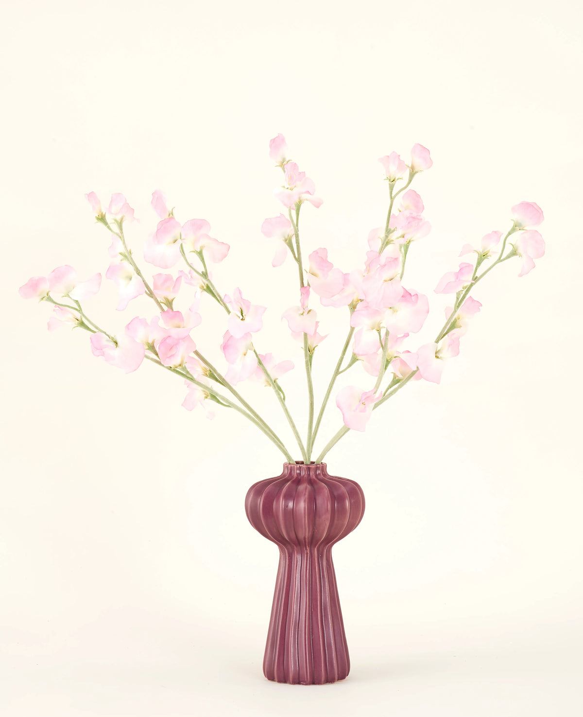 El ramo de flores artificiales de seda Pea rosa claro, con varios tallos de guisantes de color rosa claro en un jarrón acanalado de color burdeos, se alza sobre un fondo pálido: ideal para cualquier interior de flores artificiales.