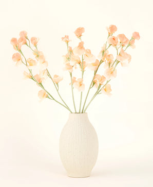 El jarrón de cerámica de color crema contiene el ramo de flores artificiales Silk Pea Peach, con delicadas flores color melocotón y rosa claro con largos tallos verdes sobre un fondo claro y neutro.