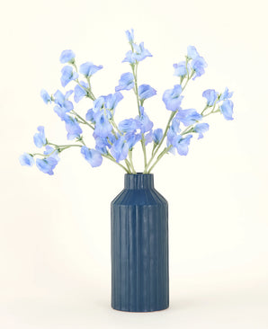 Un jarrón de cerámica acanalado azul exhibe el ramo de flores artificiales de seda Pea Blue, con sus delicadas flores artificiales de color azul claro sobre un fondo liso y claro.