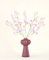 El ramo de flores artificiales de seda Lila guisante presenta tallos largos de delicadas flores de color violeta claro y hojas verdes en un jarrón de cerámica malva con crestas verticales, sobre un fondo liso y blanquecino.