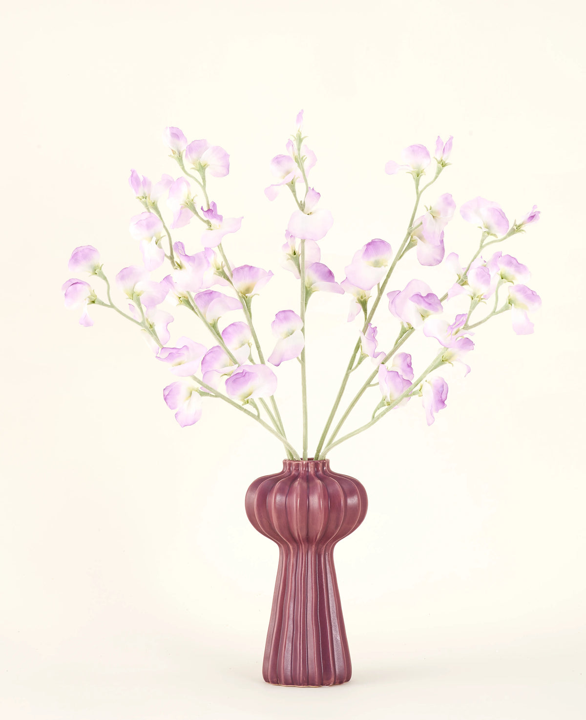 El ramo de flores artificiales de seda Lila guisante presenta tallos largos de delicadas flores de color violeta claro y hojas verdes en un jarrón de cerámica malva con crestas verticales, sobre un fondo liso y blanquecino.