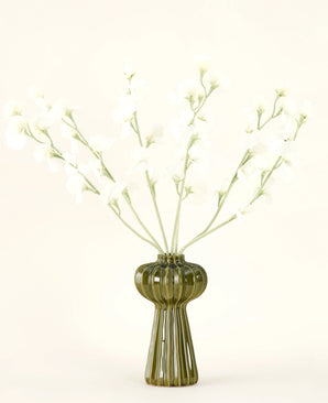 Un jarrón de vidrio acanalado verde sostiene el ramo de flores artificiales de seda color crema guisante con delicadas flores de seda blanca y tallos delgados, sobre un fondo liso blanquecino.