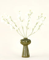 Un jarrón de vidrio acanalado verde sostiene el ramo de flores artificiales de seda color crema guisante con delicadas flores de seda blanca y tallos delgados, sobre un fondo liso blanquecino.
