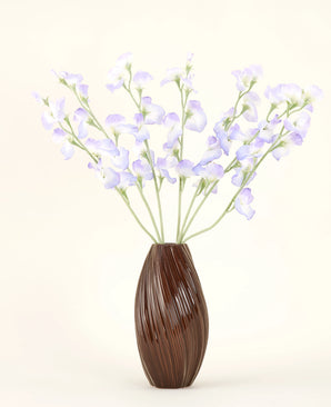 Un jarrón marrón acanalado sostiene el ramo de flores de seda artificial Pea Lavender, con delicados pétalos y tallos verdes sobre un fondo blanquecino liso para crear una atmósfera sutil de cabaña.