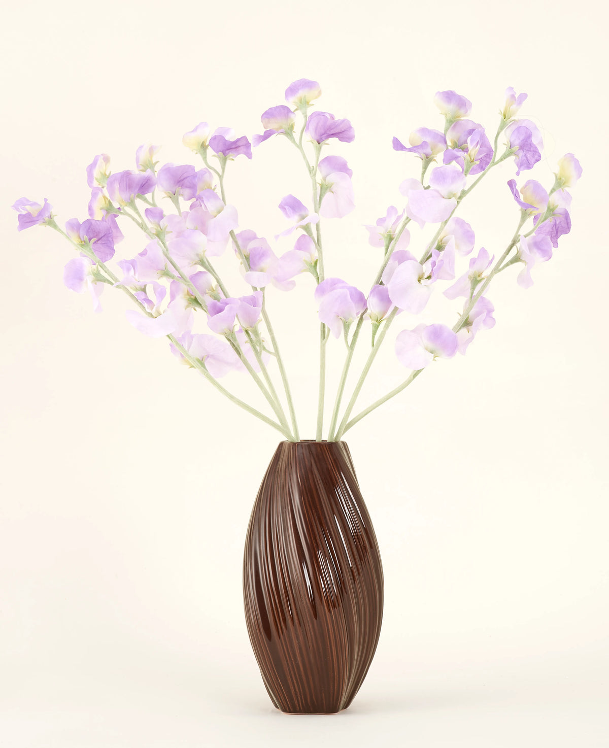 Un jarrón marrón texturizado sostiene el ramo de flores de seda artificial, flores de seda de color violeta claro con tallos verdosos sobre un fondo liso y claro.