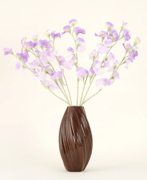Un jarrón marrón texturizado sostiene el ramo de flores de seda artificial, flores de seda de color violeta claro con tallos verdosos sobre un fondo liso y claro.