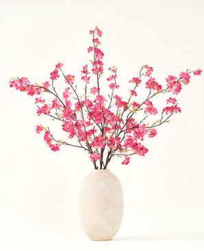 El ramo de flores artificiales de seda Flor de cerezo fucsia de 150 cm presenta vibrantes tallos de flores de cerezo fucsia con hojas verdes en un sencillo jarrón blanco, que complementará cualquier interior sobre un fondo claro.