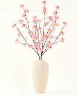 Un jarrón alto de cerámica beige contiene varias ramas del ramo de flores artificiales Silk Flores rosas, con delicadas flores rosas y hojas verdes, sobre un fondo blanco liso.