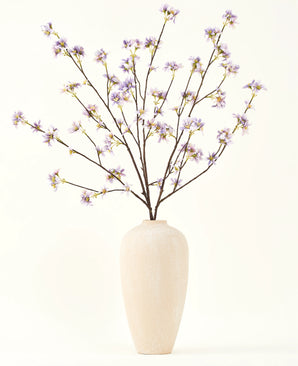 Un jarrón alto de cerámica de color crema contiene varias ramas delgadas con el texto Ramo de flores artificiales de seda Rama de flores artificiales de color púrpura, sobre un fondo liso y claro.
